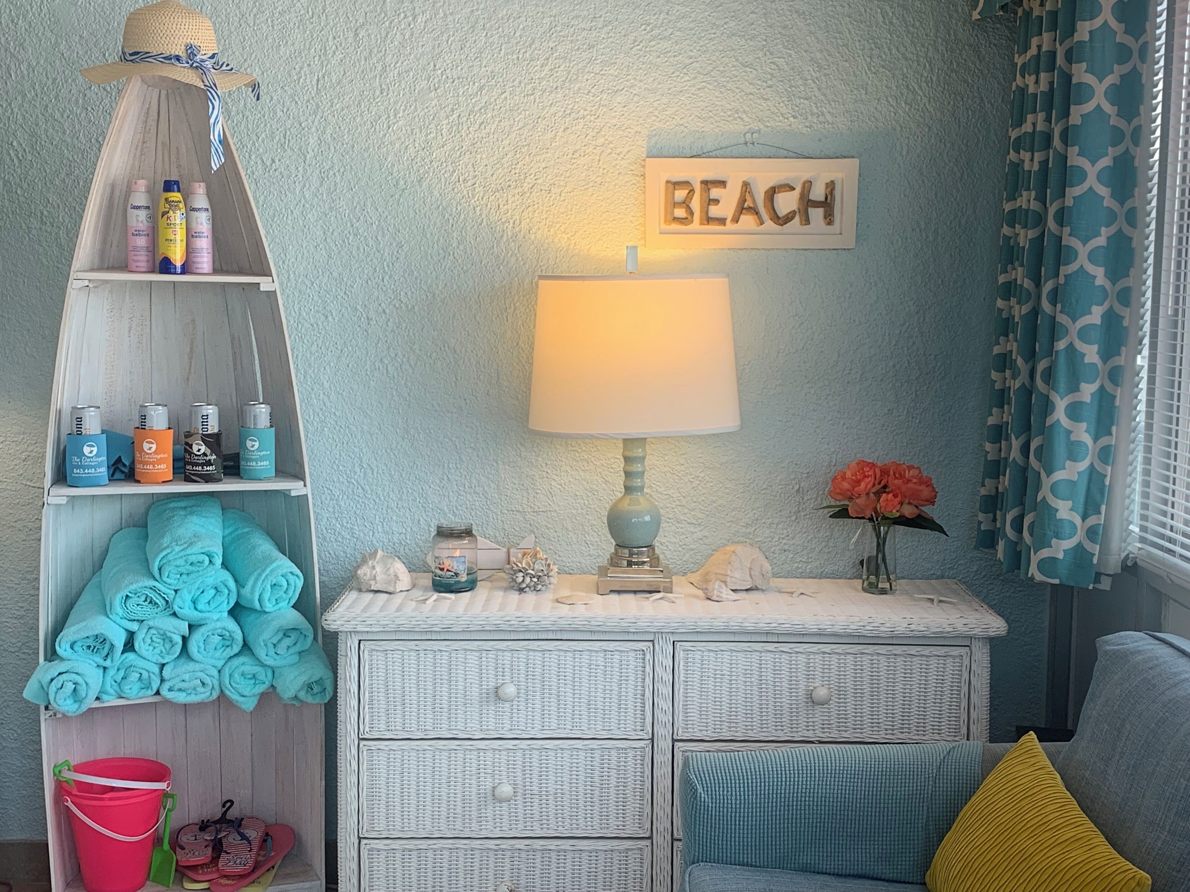 A beach-themed room with a wicker dresser, a shelf with baby products, a beach sign, a lamp, and a window with patterned curtains.