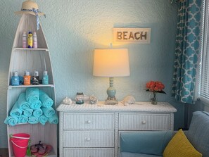 A beach-themed room with a wicker dresser, a shelf with baby products, a beach sign, a lamp, and a window with patterned curtains.