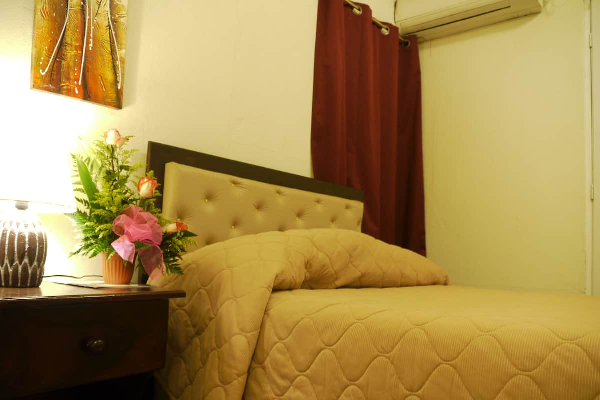 A neatly made bed with a quilted headboard, a bedside table with a lamp and a potted plant, and a red curtain.