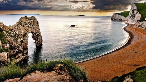 Un paisaje costero con una imponente formación rocosa en arco, una playa de arena y acantilados.
