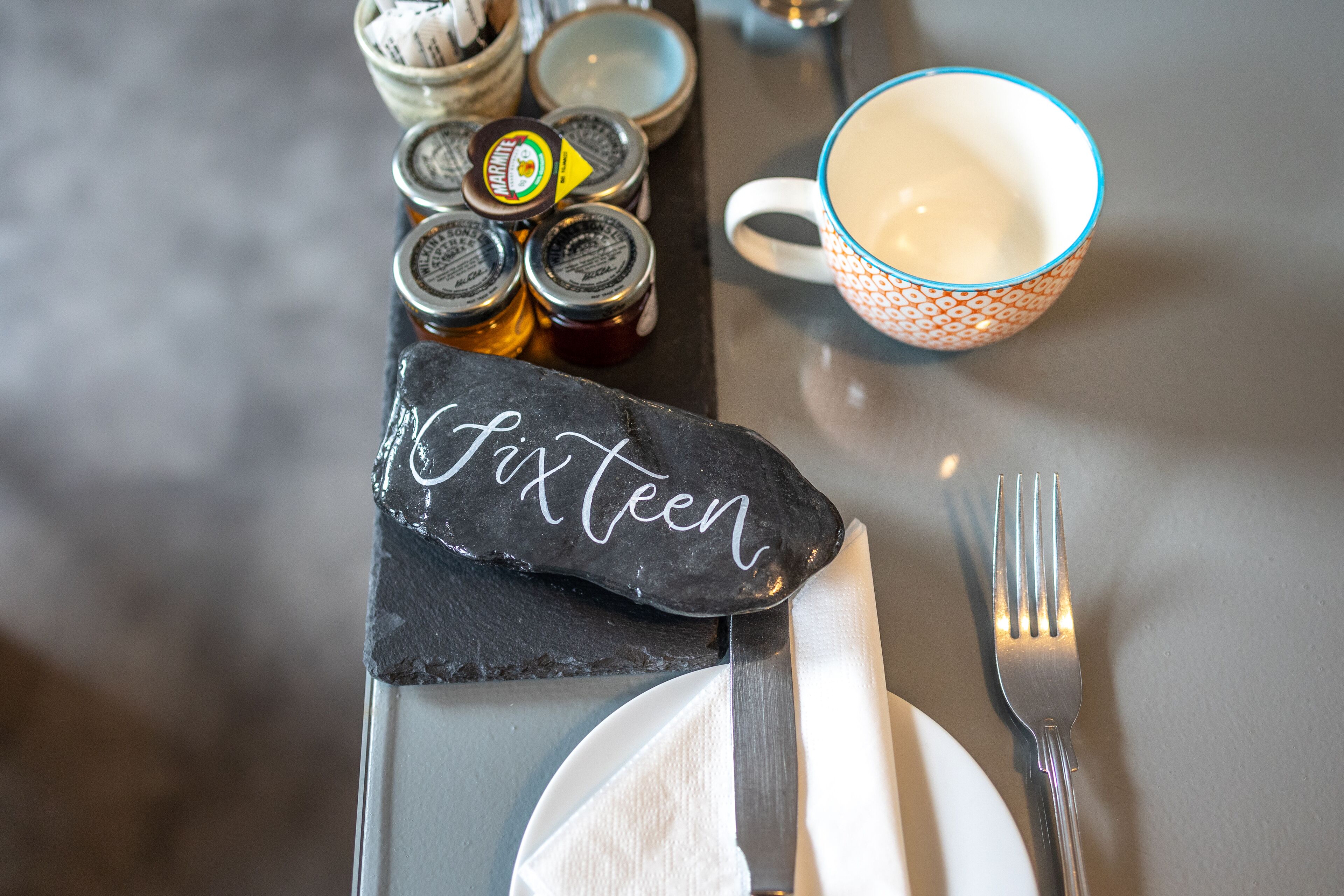 A table set with a slate napkin holder labeled "Fifteen," a white napkin, a fork, a knife, and a patterned mug.