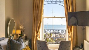 Habitación de hotel con una cama, dos sillones, una mesita, un televisor, una ventana con vista al mar y un balcón con mesa y sillas.