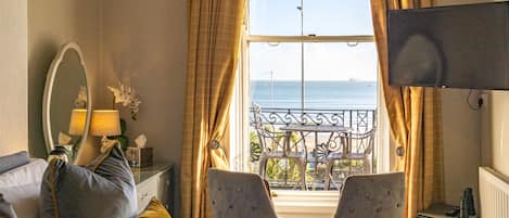 A hotel room with a bed, two armchairs, a small table, a TV, a window with a view of the sea, and a balcony with a table and chairs.