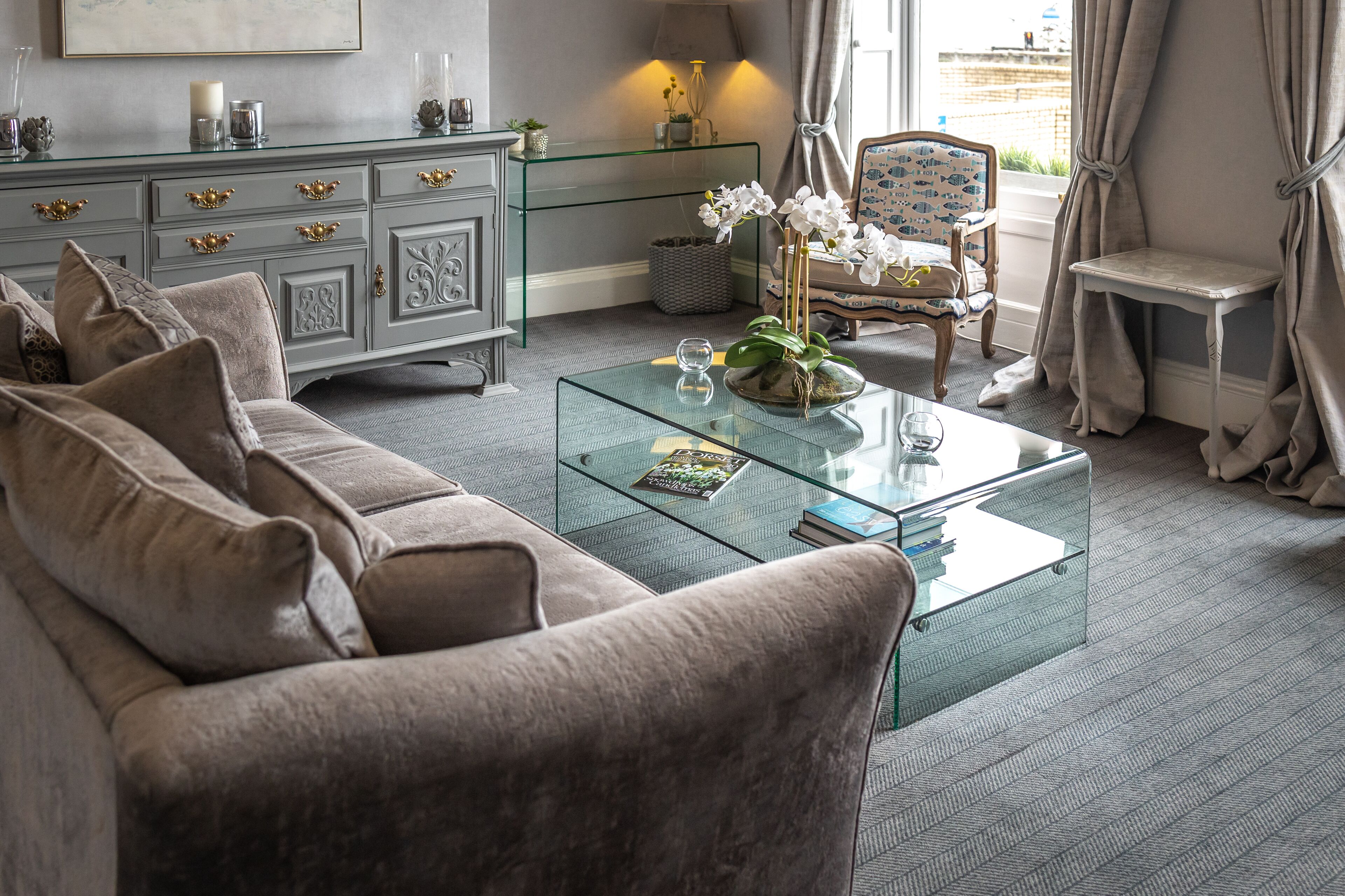 A modern living room with a glass coffee table, grey sofa, and a decorative chair.