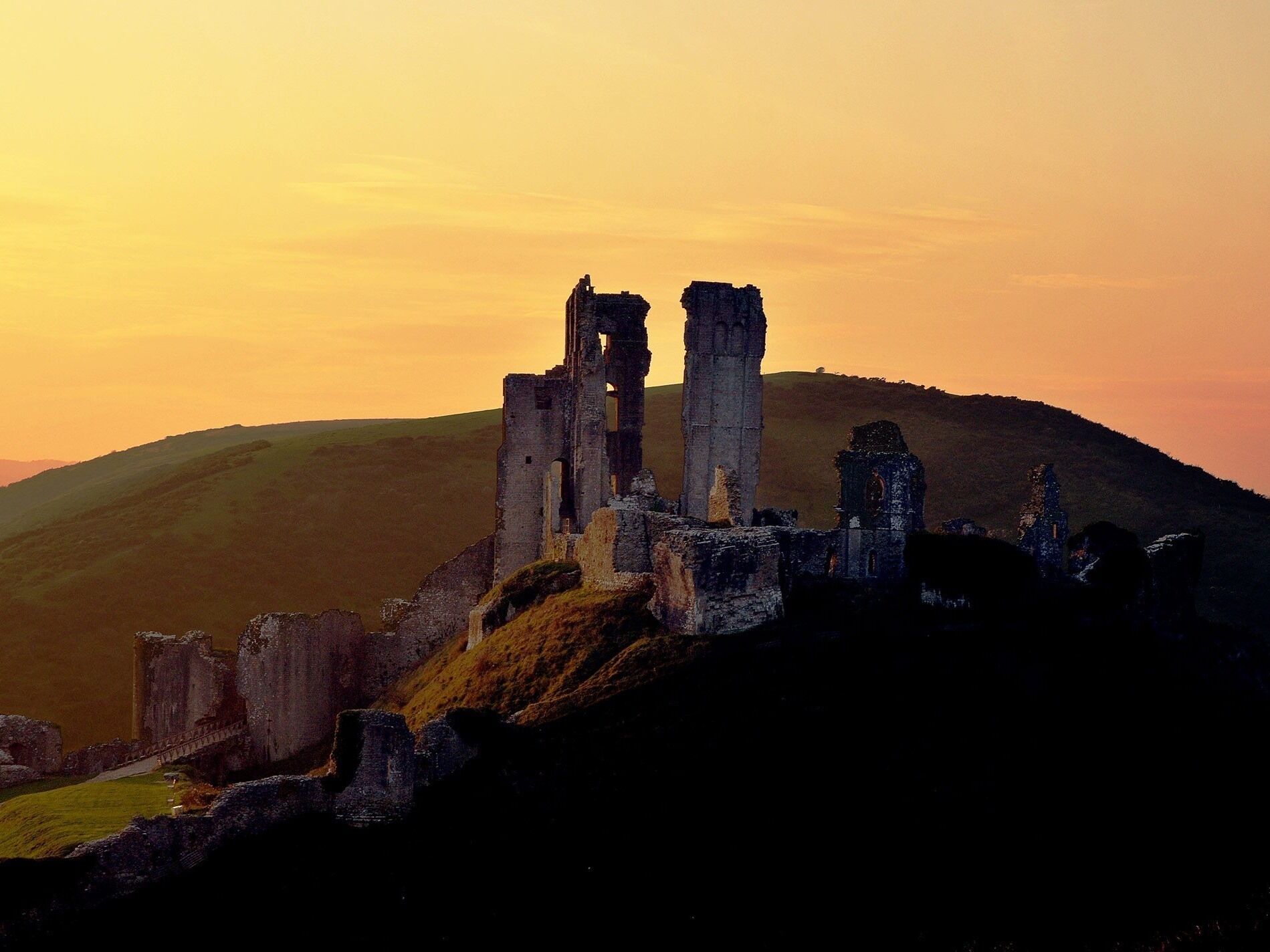 A historic castle ruin with multiple towers and walls, set against a hilly backdrop during sunset.