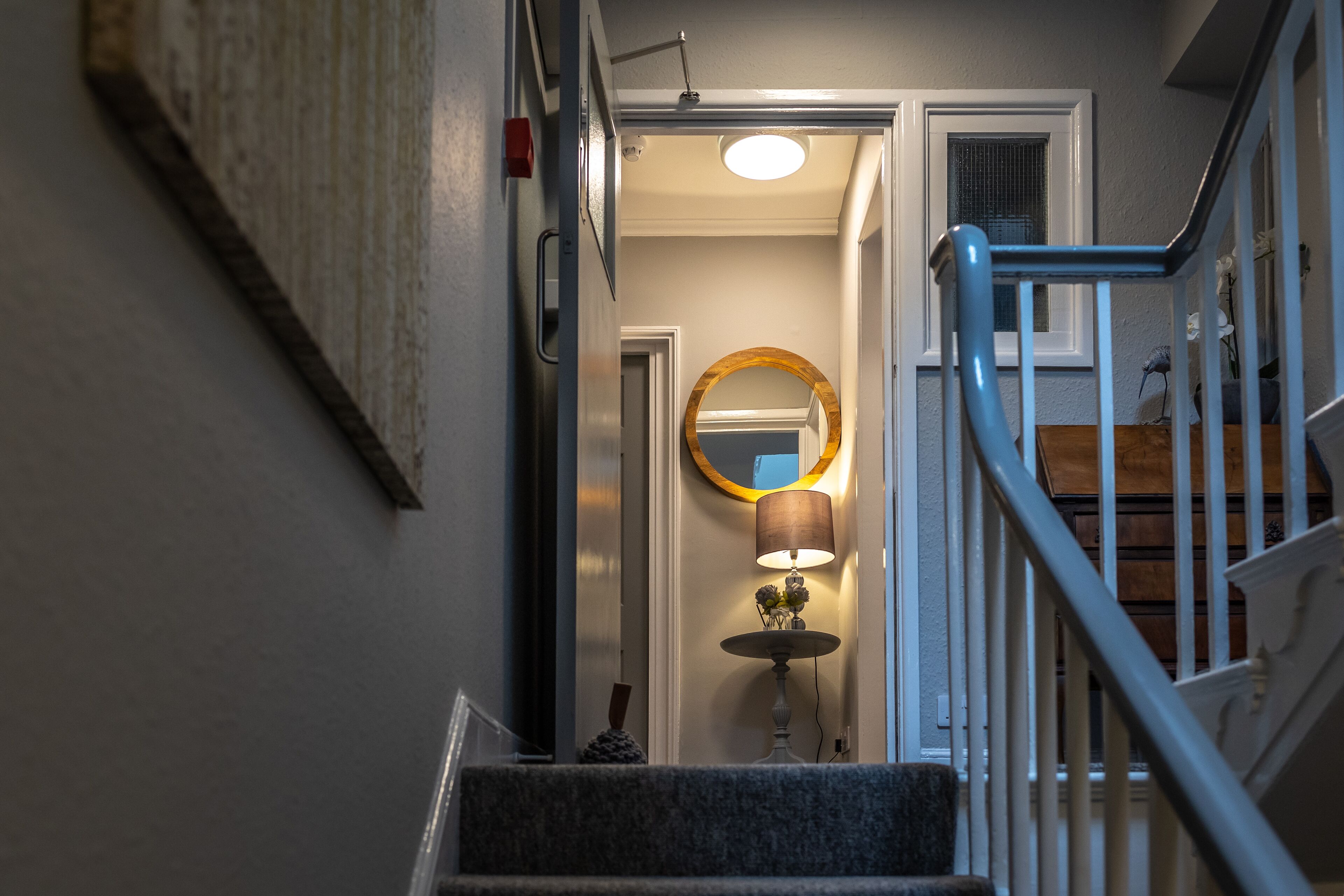A staircase with a blue handrail leading to a landing where a round mirror hangs on the wall, a table lamp, and a small decorative item.