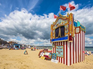A beachside carnival tent with a striped red and white exterior, a sign for the Punch & Judy show, and a beach area with people and beach chairs. - Gloucester House (Weymouth)