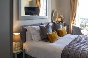 A neatly made bed with a gray headboard, white and gray pillows, and a mustard-colored throw. A small bedside table with a lamp and a mirror is visible. - Gloucester House (Weymouth)