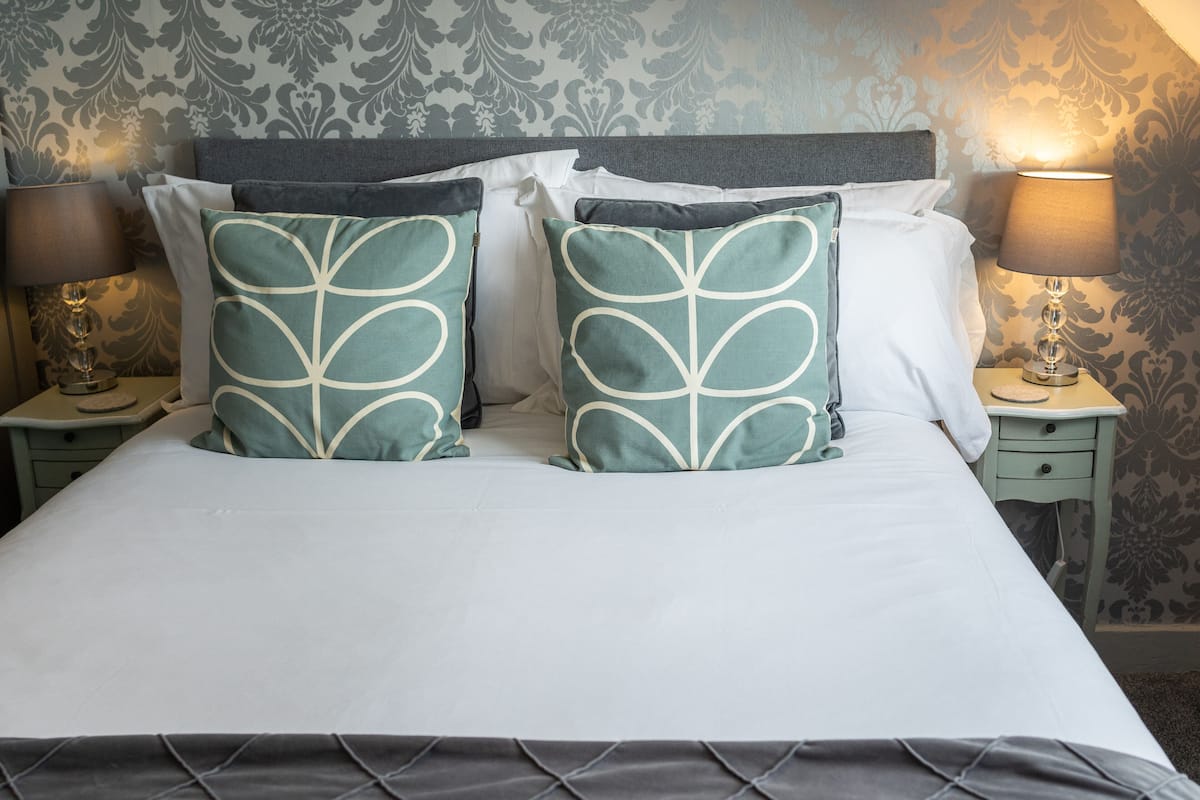Una cama bien tendida con dos almohadones decorativos, un cabecero y dos lámparas de noche a cada lado.