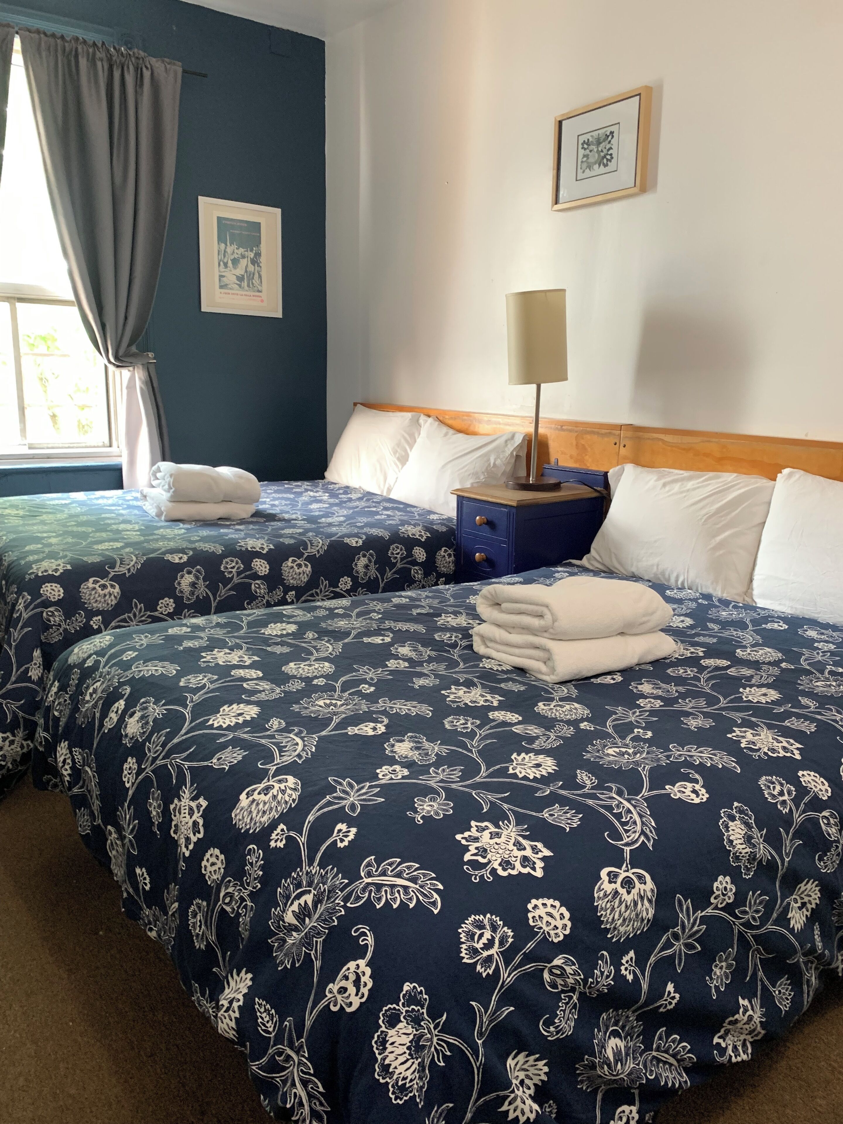 A hotel room with two beds, each with a floral-patterned bedspread, a bedside table with a lamp, and a window with curtains.