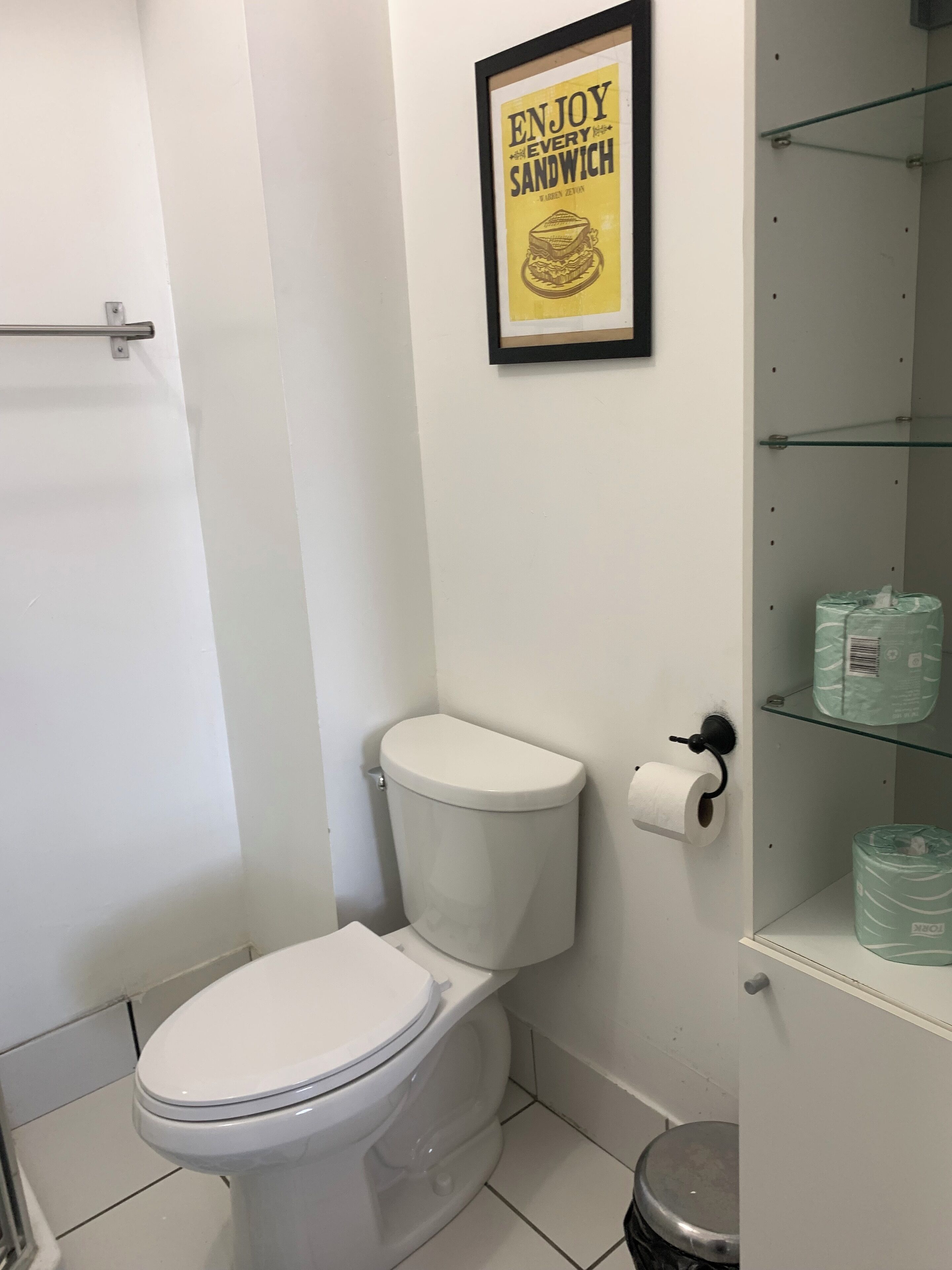 Un baño con inodoro, un cartel enmarcado sobre el inodoro y un estante con papel higiénico y un cubo de basura.