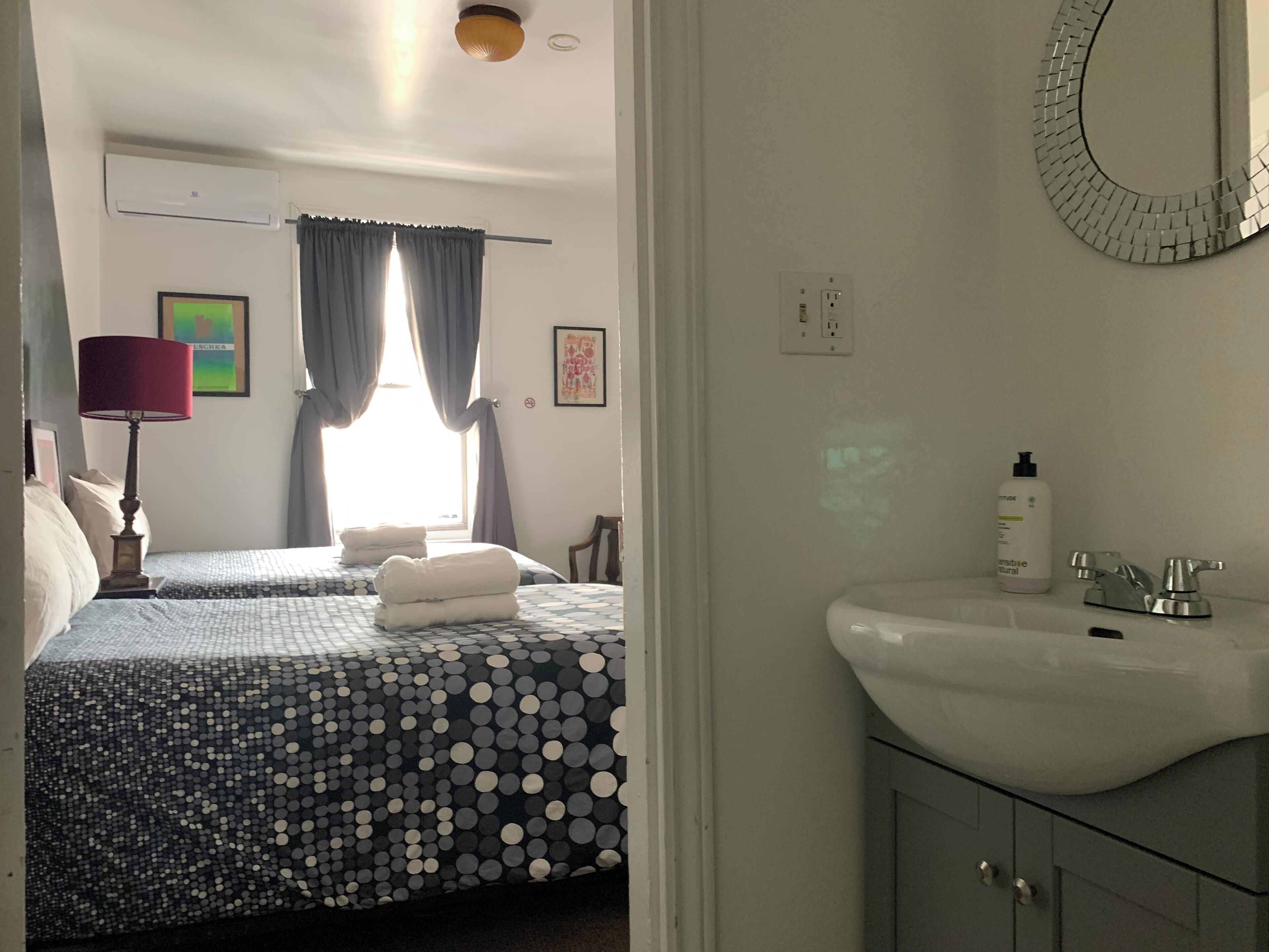 Une chambre d’hôtel avec deux lits, une salle de bain avec un lavabo et un miroir, et une fenêtre avec des rideaux.