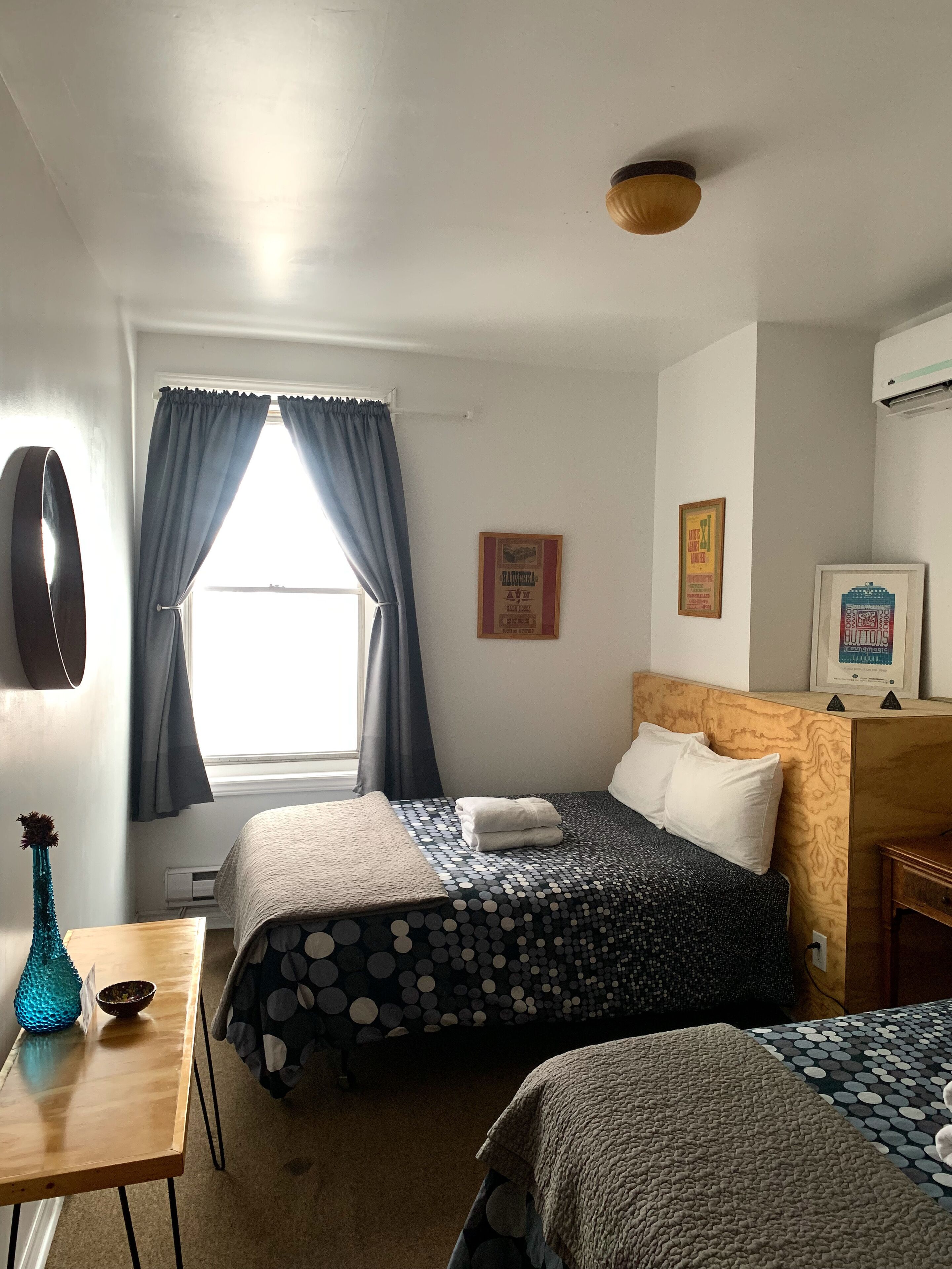 A hotel room with two beds, a wooden desk, a blue vase, and a window with curtains.
