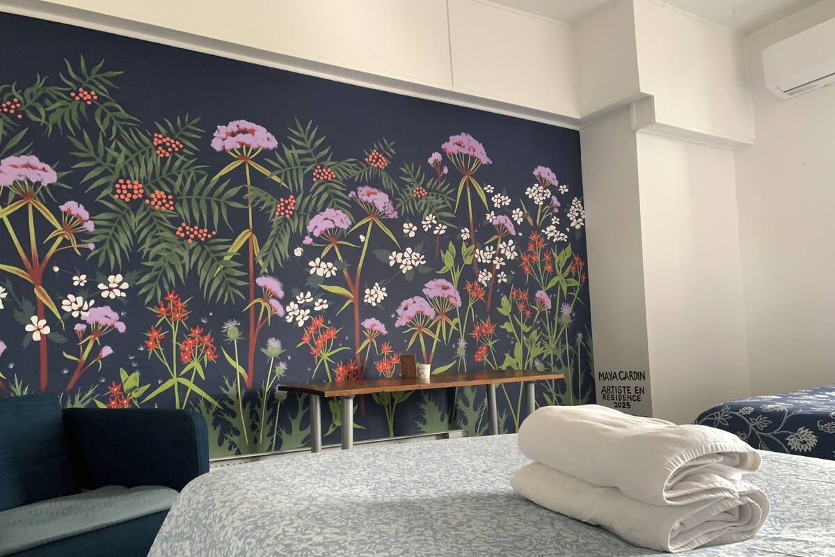 A hotel room with a floral mural, a bed with a patterned bedspread, a nightstand, and a blue chair.