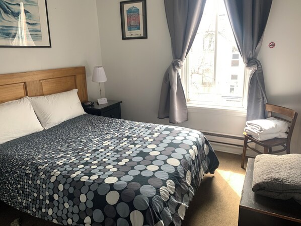 A neatly made bed with a polka dot bedspread, a wooden headboard, a bedside table with a lamp, a chair with a folded towel, and a window with curtains. - Pensione Popolo (Montreal)