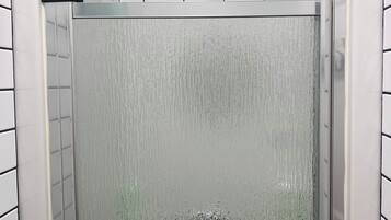 Ducha con puerta de vidrio, paredes de azulejos blancos y ducha superior.