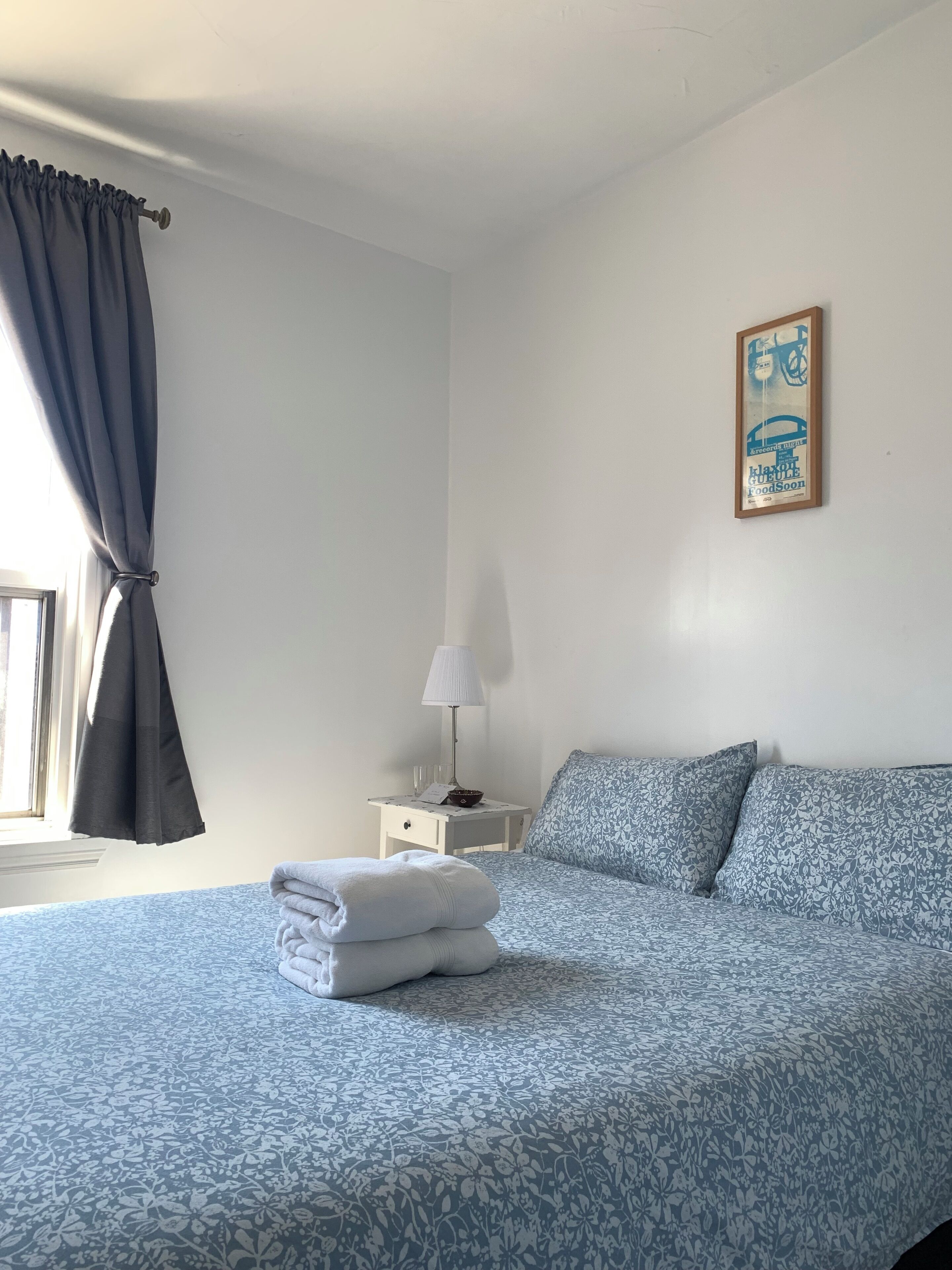 Una cama bien hecha con estampado floral, una mesita de noche con una lámpara y un cuenco, y un póster enmarcado en la pared.