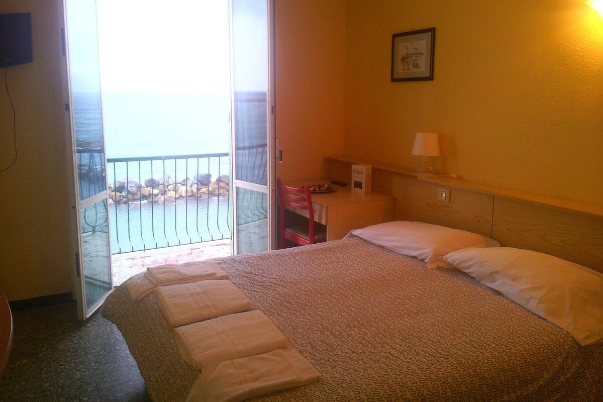 A hotel room with a bed, a desk, a chair, a lamp, and a view of the sea through a balcony door.