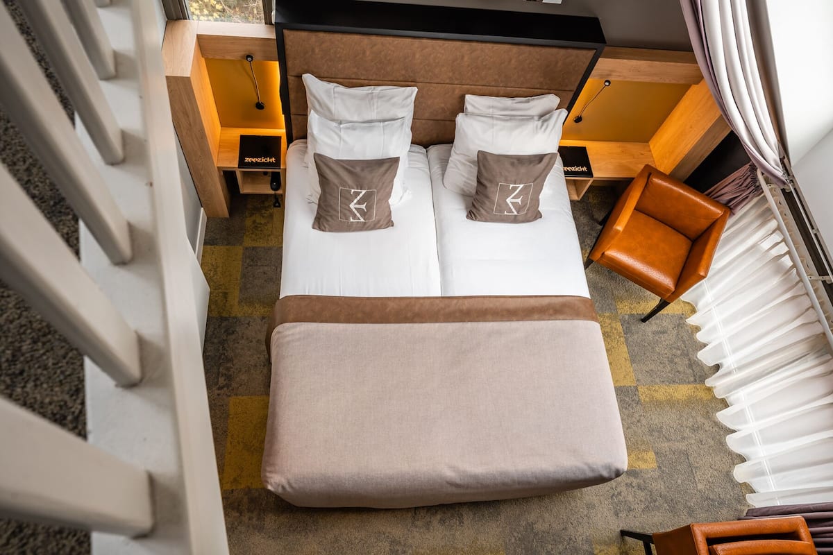 A neatly arranged bed with pillows, a bedside table, and a chair in a room with a checkered carpet.