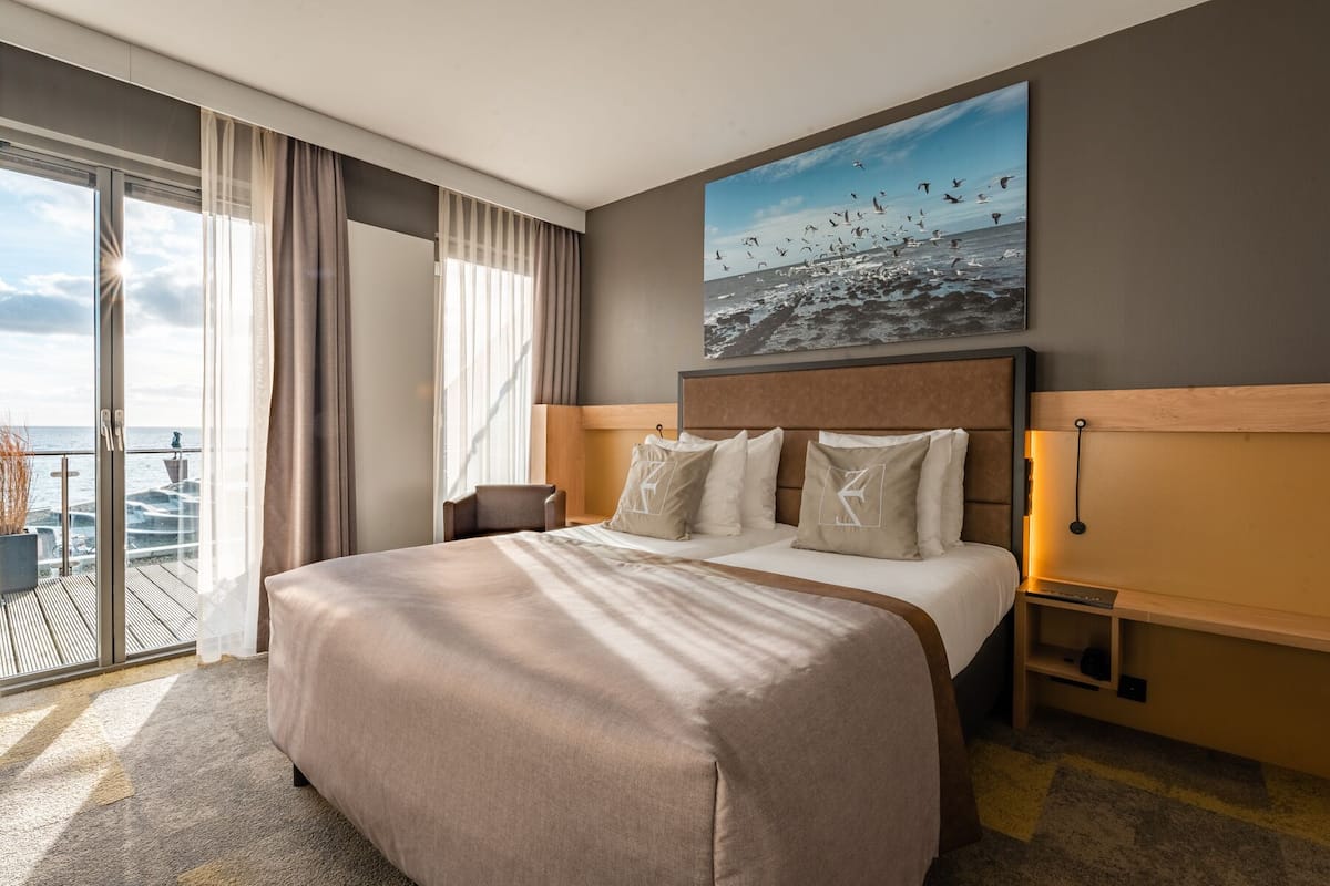 A hotel room with a large bed, a view of the sea, and a painting of birds on the wall.
