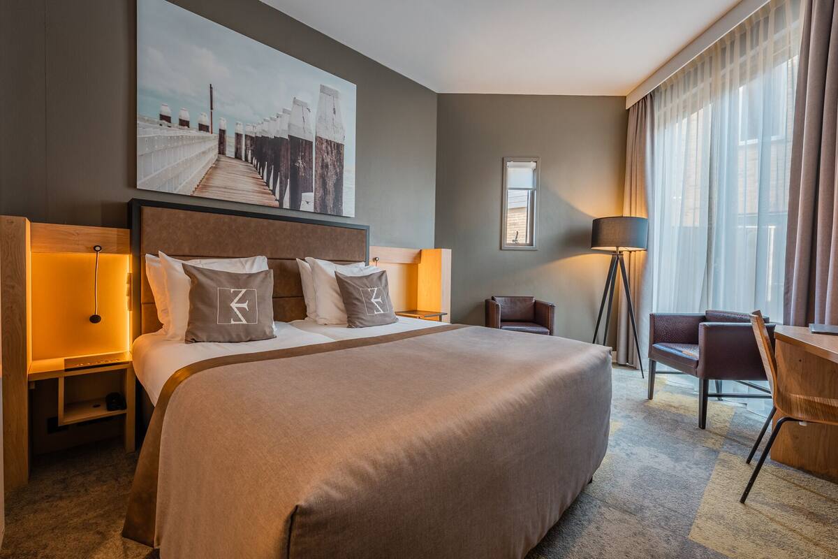 A hotel room with a large bed, a desk, a chair, a lamp, and a painting on the wall.
