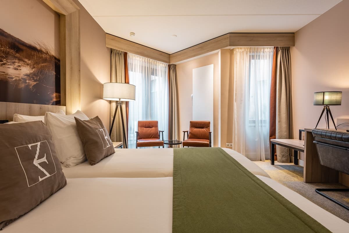 A hotel room with two beds, a desk, a chair, and a lamp.