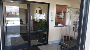 A hotel entrance with a glass door, a reception desk, a potted plant, a metal cart, and a wall-mounted phone. - Briarwood Inn (Fairfield)