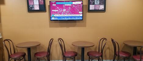L’intérieur d’un restaurant présente six tables et chaises en bois, un téléviseur fixé au mur qui affiche les prévisions météorologiques, et des œuvres d’art encadrées qui décorent le mur.