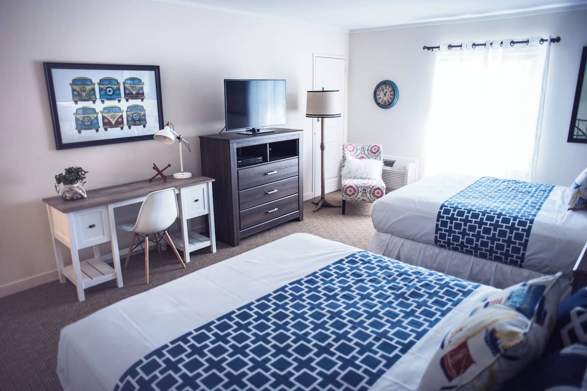 Habitación de hotel con dos camas, un escritorio con silla, televisor, lámpara, una silla con cojín estampado y un reloj de pared.