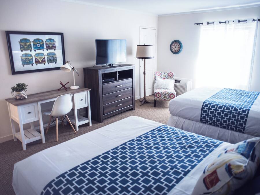 Habitación de hotel con dos camas, un escritorio con silla, televisor, lámpara, una silla con cojín estampado y un reloj de pared.