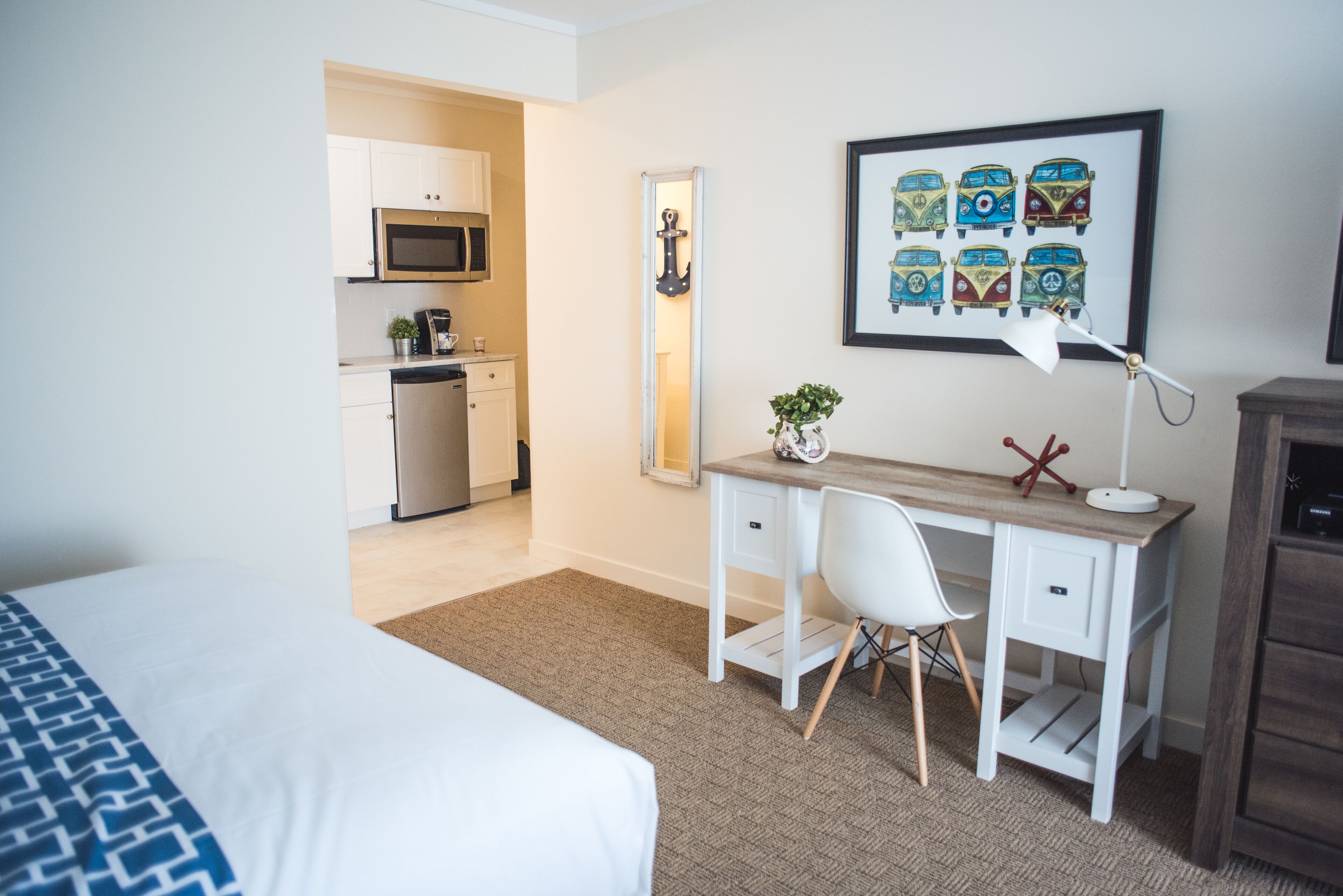 Quarto de hotel com cama, escrivaninha com cadeira, uma pequena planta, luminária de mesa, quadro com carros antigos e área de cozinha com micro-ondas e armários.
