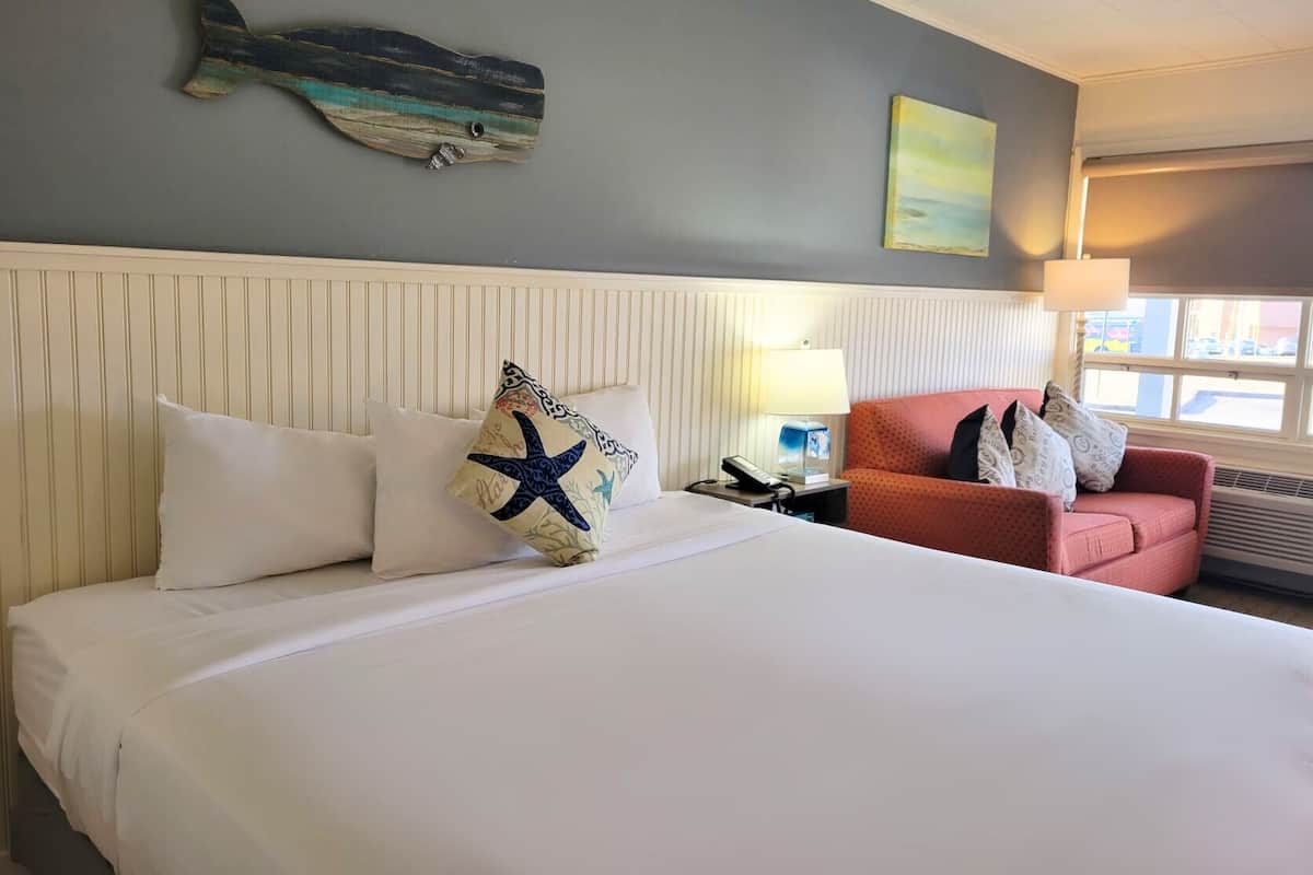 Habitación de hotel con una cama, un sillón rojo, una mesita de noche, una lámpara y un adorno en la pared.