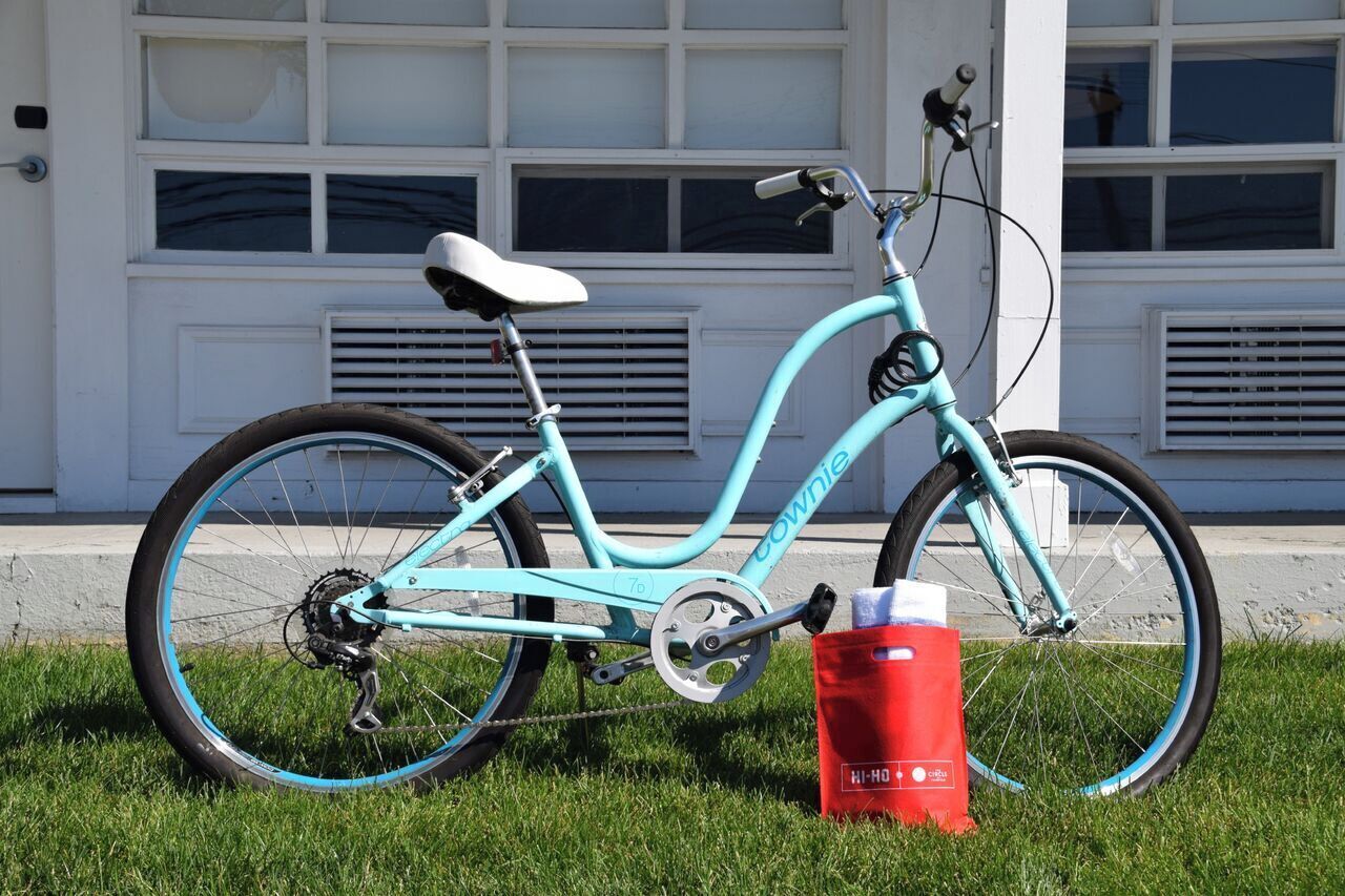 A turquoise bicycle with a white seat and 'COWNIE' branding, parked on grass in front of a white building with large windows and vents.