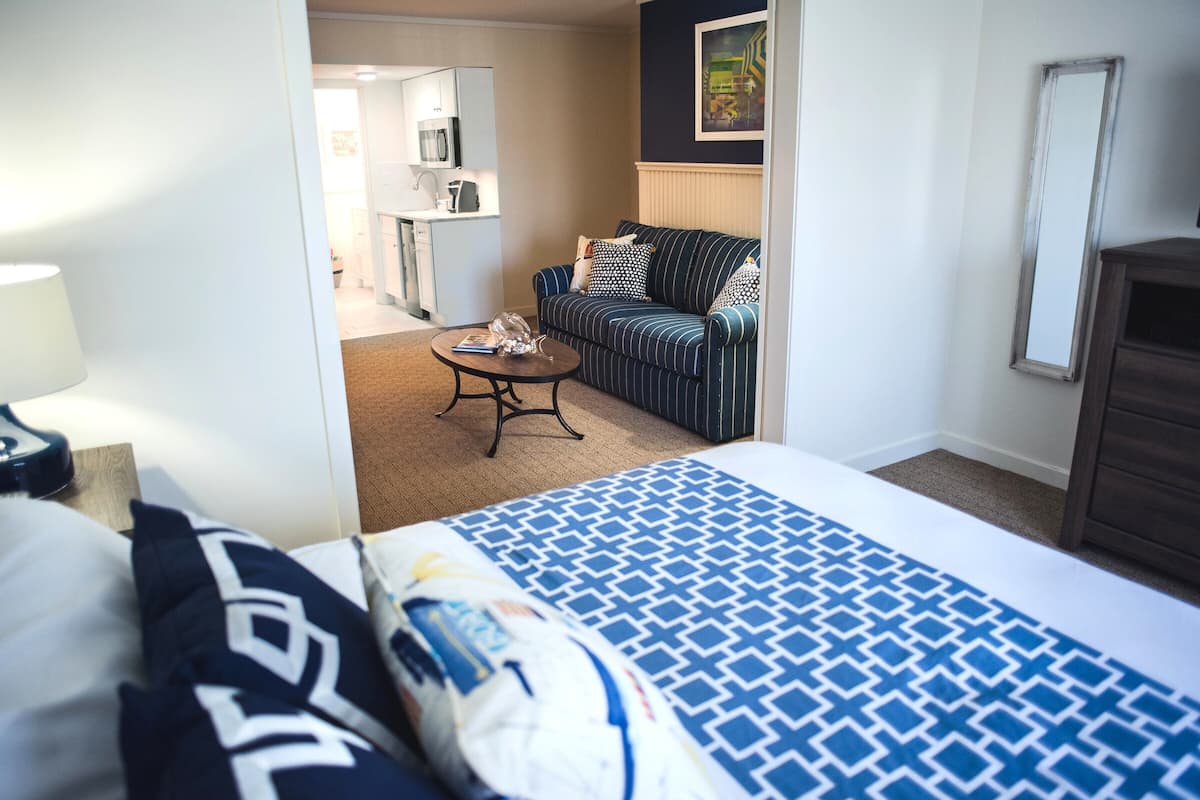 Habitación de hotel con cama, sofá, mesa de centro y una cocina visible a través de una puerta abierta.
