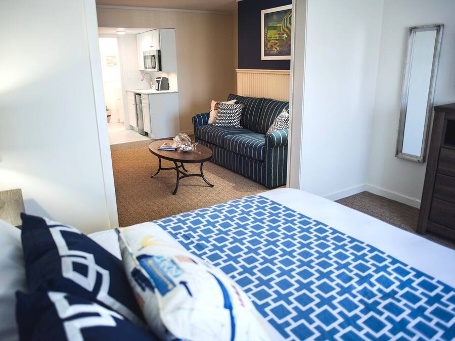 Et hotelværelse med seng, sofa, sofabord og et køkkenområde, der kan ses igennem en åben dør.