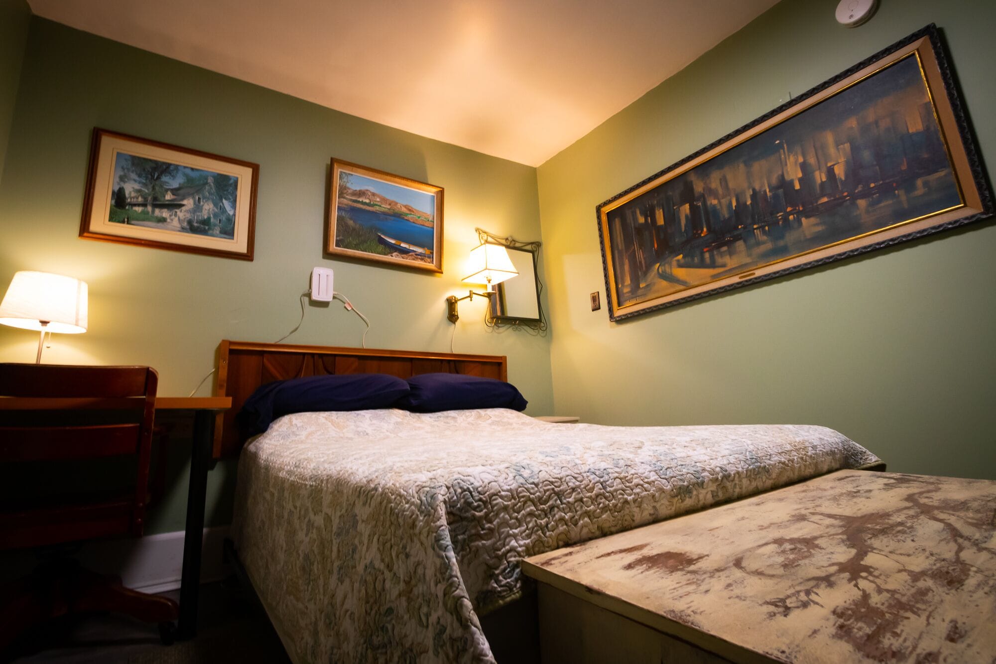 A hotel room with a bed, a chair, a desk, and a lamp. There are three framed paintings on the wall.