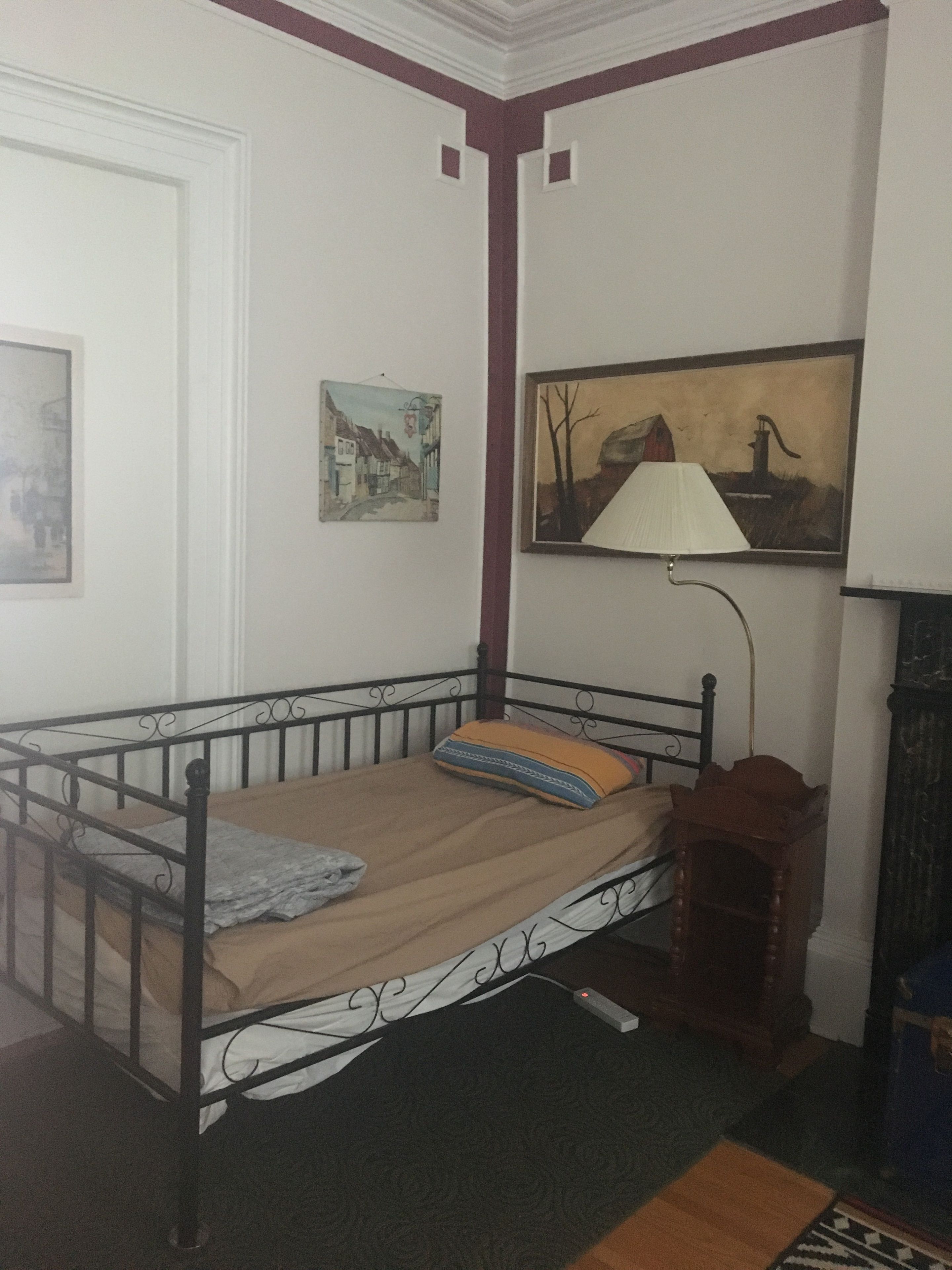 A single bed with a metal frame, a wooden headboard, and a patterned bedspread. There is a small bedside table with a lamp and a picture frame on the wall above the bed.