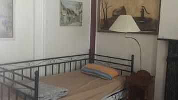 A single bed with a metal frame, a wooden headboard, and a patterned bedspread. There is a small bedside table with a lamp and a picture frame on the wall above the bed.