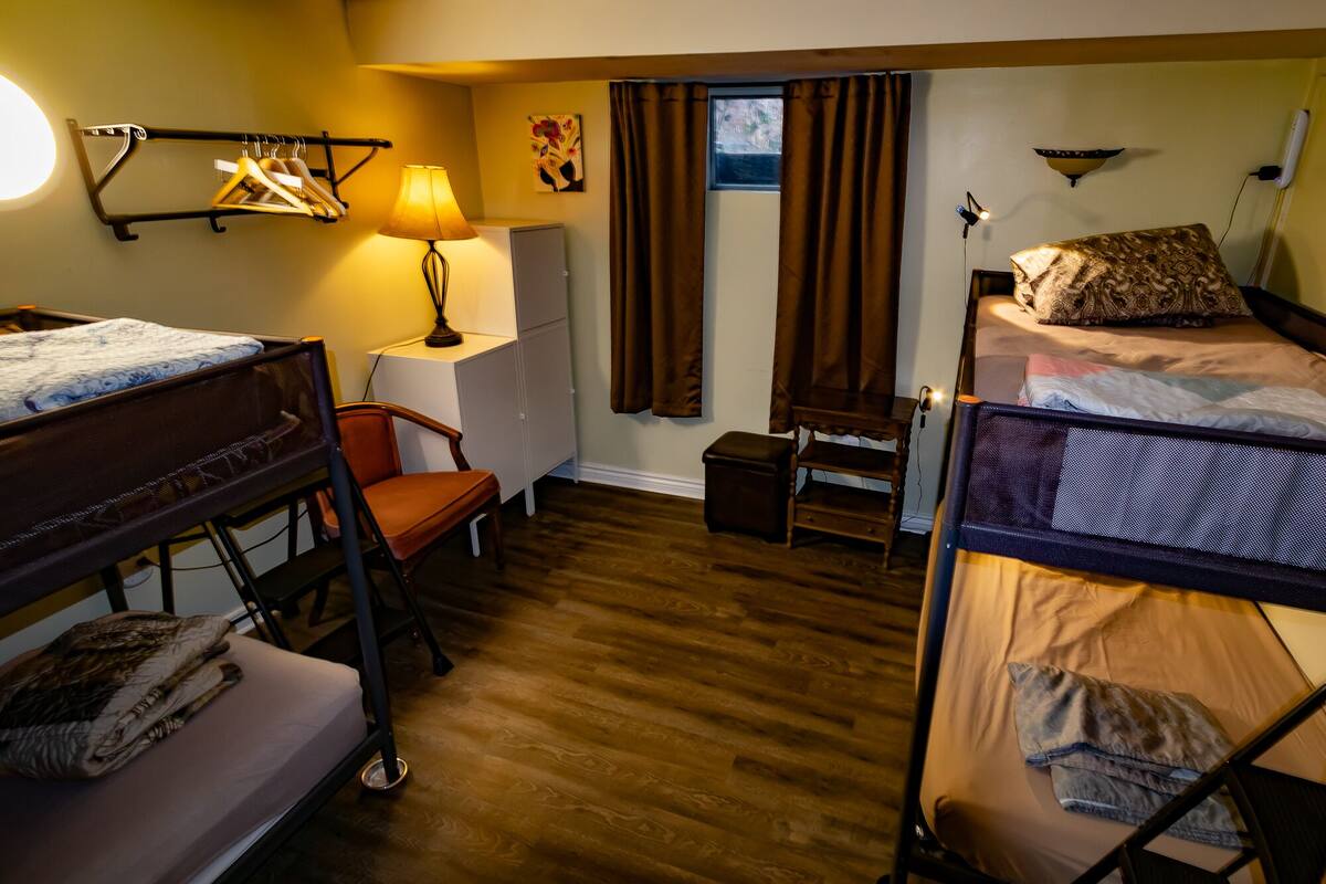 A compact hotel room with two bunk beds, a desk with a lamp, a chair, a small table, and a window with curtains.