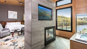 A modern hotel room with a fireplace, a large TV mounted on a stone wall, a bed with white linens, a sitting area with a sofa and armchairs, and a view of a lake through large windows. - Beaver Lakefront Cabins - Couples Only Getaways (Eureka Springs)