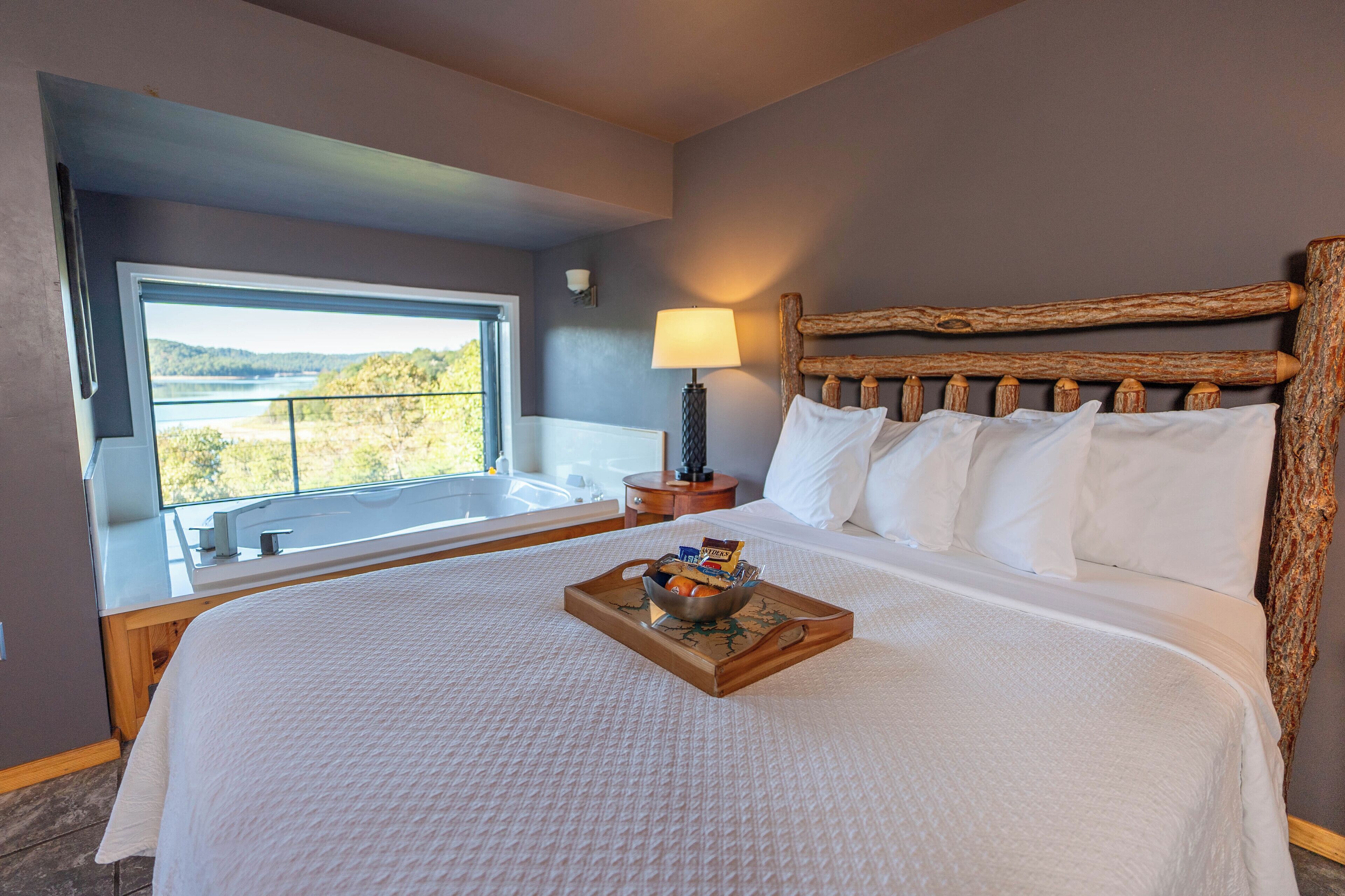 Un dormitorio con una cama grande, cabecera de madera, ventana con vistas, mesita de noche con lámpara y bandeja con artículos para el desayuno.