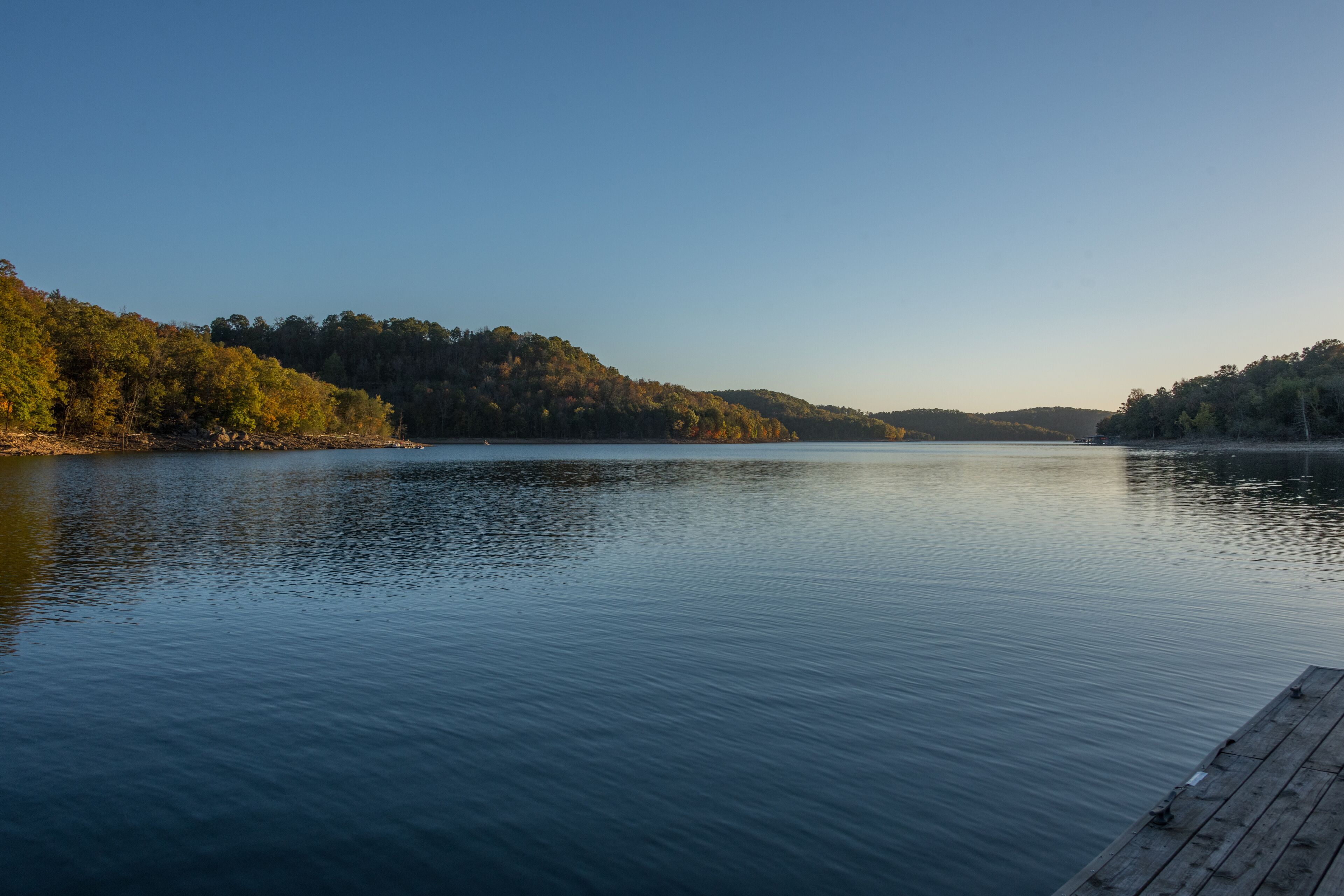 A calm lake with a wooden dock extending into the water, surrounded by forested hills.