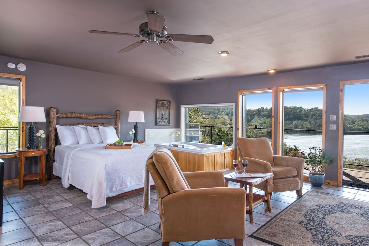Une chambre à coucher avec un lit, deux chaises, une table, un ventilateur de plafond et une vue sur un lac.