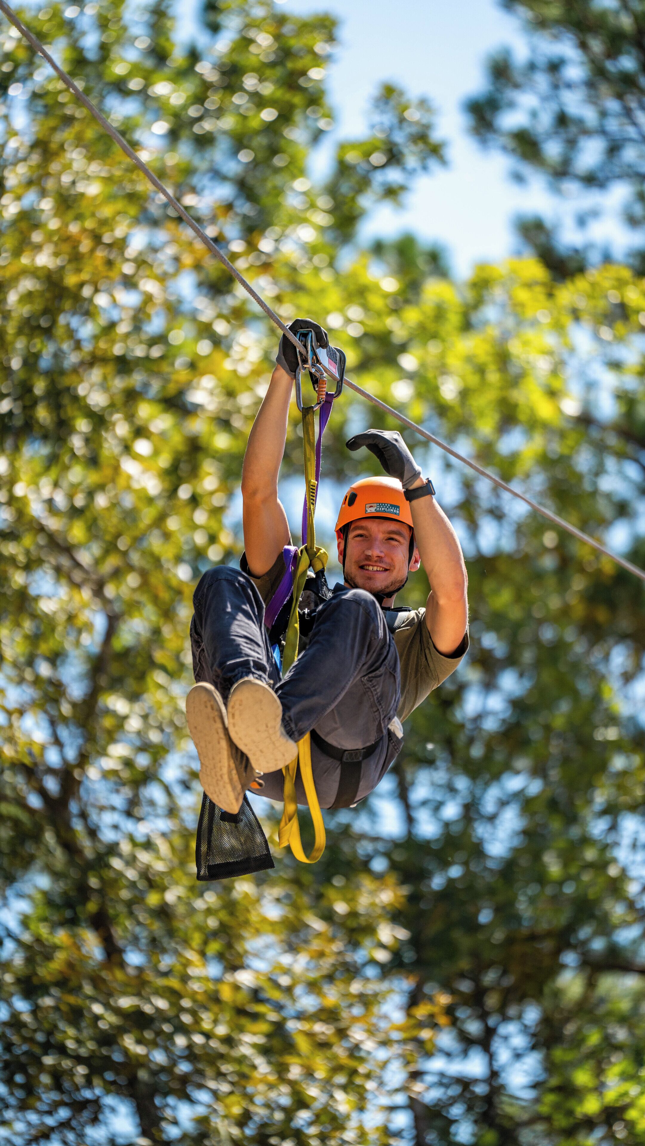 A person engaged in a zip-lining activity, suspended in mid-air with a harness and helmet, against a backdrop of trees.