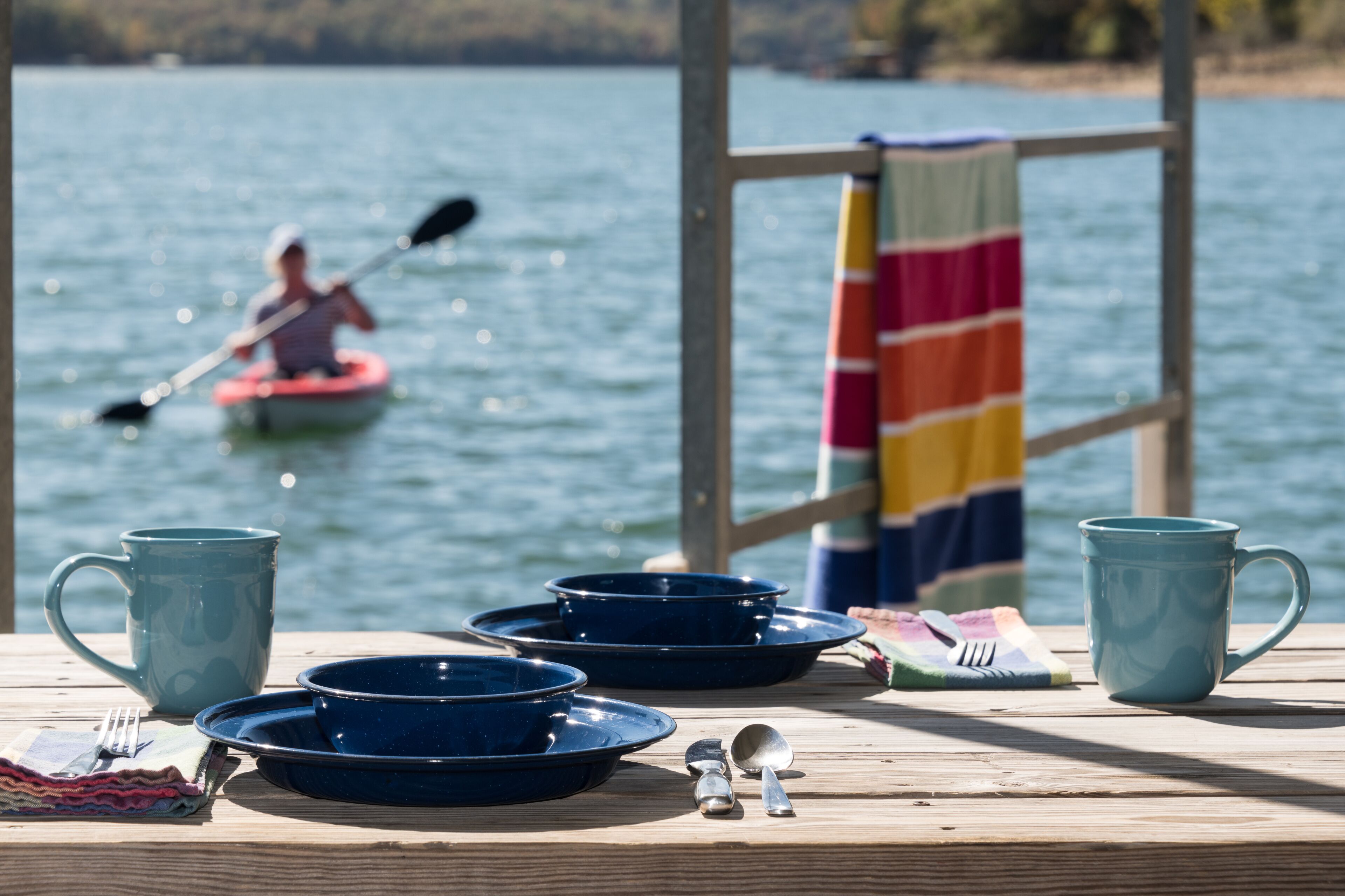 A wooden table with blue dishes, a mug, and utensils, set against a lake view with a person kayaking in the background.