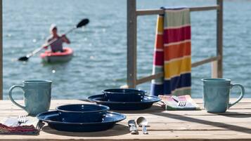 A wooden table with blue dishes, a mug, and utensils, set against a lake view with a person kayaking in the background.