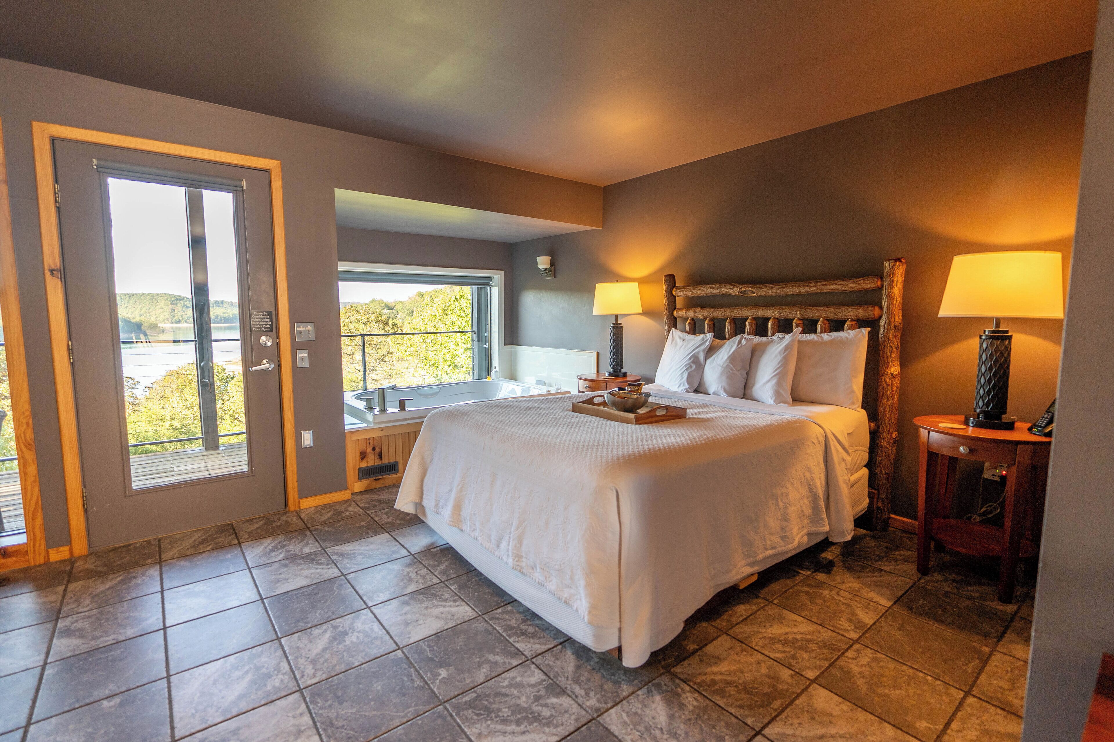 A bedroom with a large bed, a window with a view, a bedside table with a lamp, and a door leading to an outdoor area.