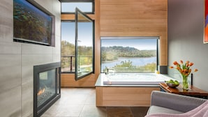 A modern bathroom with a fireplace, a large window with a view of a lake, a bathtub, and a wooden vanity with a vase of flowers. - Beaver Lakefront Cabins - Couples Only Getaways (Eureka Springs)