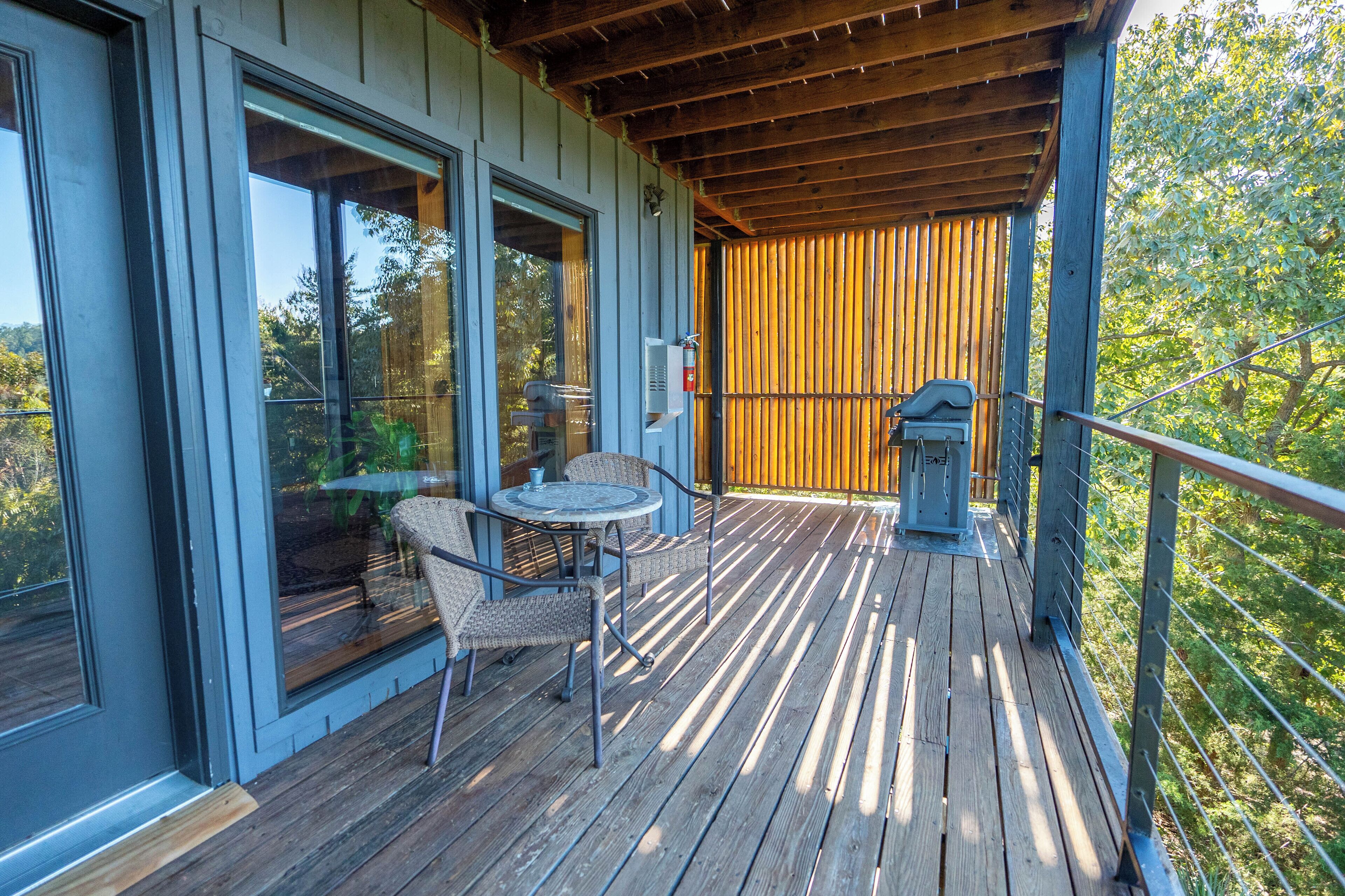 Een houten terras met een tafel, stoelen, een barbecue en uitzicht op bomen.