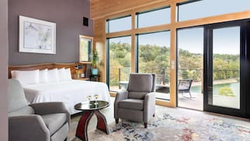 Un dormitorio moderno con una cama grande, un sillón gris, una mesita con vasos y vistas a un bosque a través de amplios ventanales.